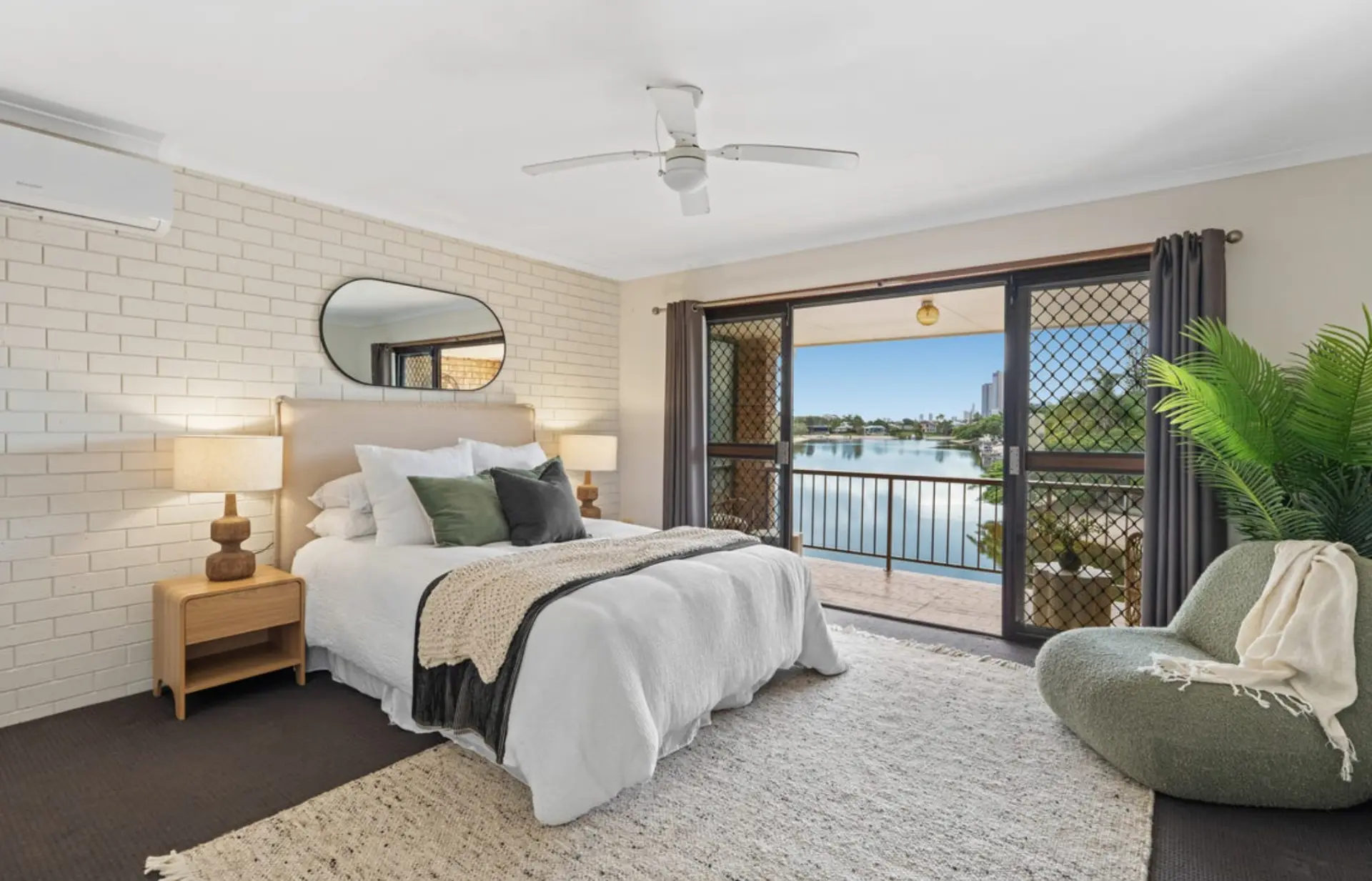 Master bedroom styled with white linen, olive cushions and a bouclé chair overlooking the water